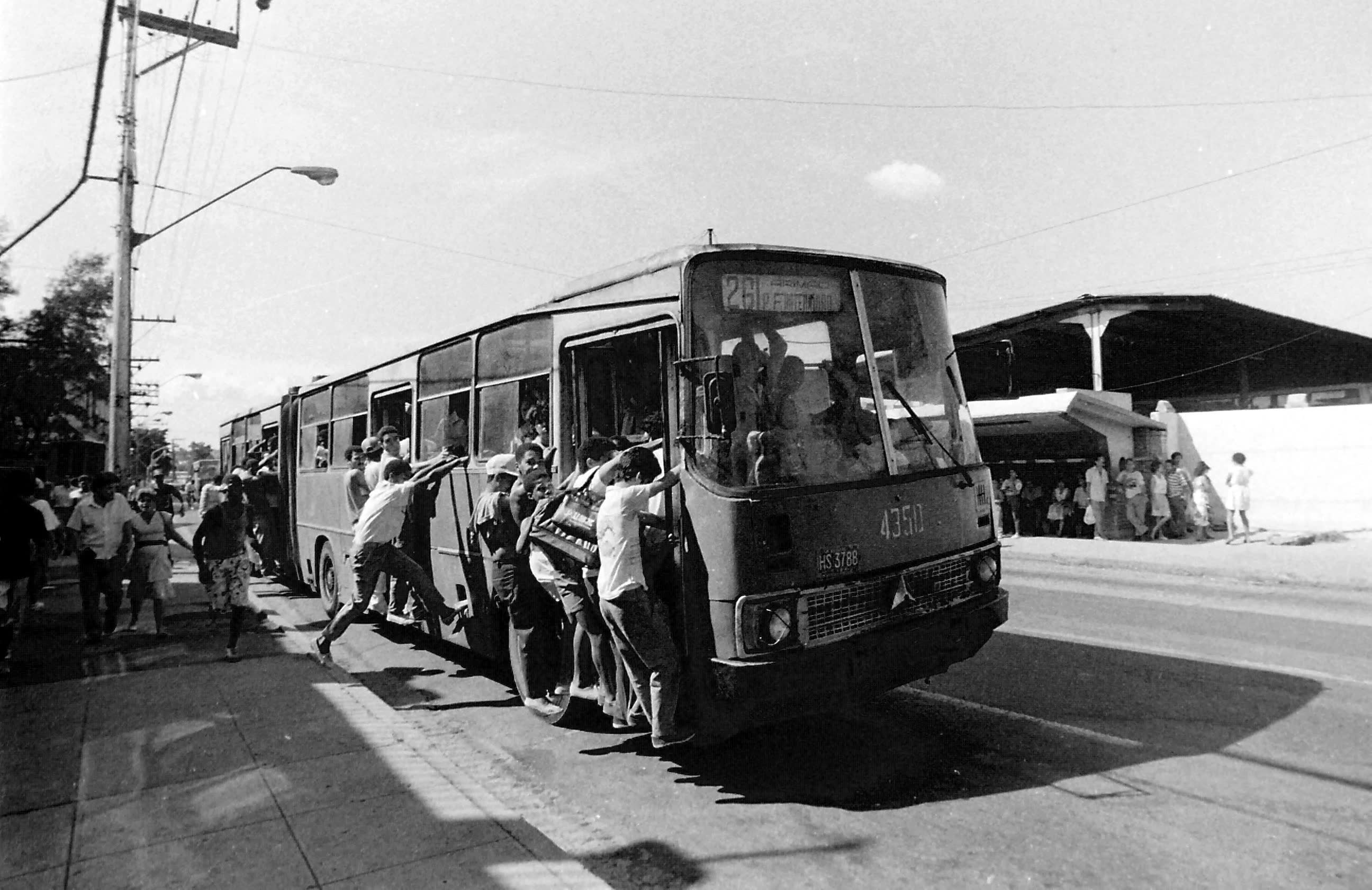 Un grupo de personas empuja un ómnibus averiado por una calle de Cuba; en blanco y negro, con más gente esperando bajo el sol